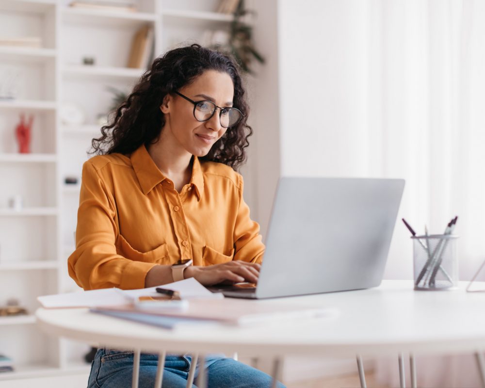Middle Eastern Lady Using Laptop Working Online Wearing Eyewear Sitting At Workplace In Modern Office. Remote Job, Technology And Career Profession Concept. Side View